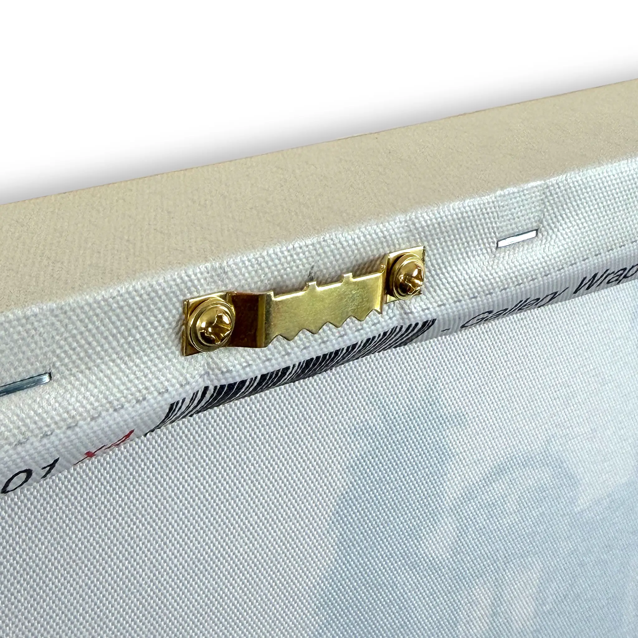 Close-up of a canvas or fabric panel showing the wooden stretcher bar, brass hanging hardware, and textured off-white fabric surface.