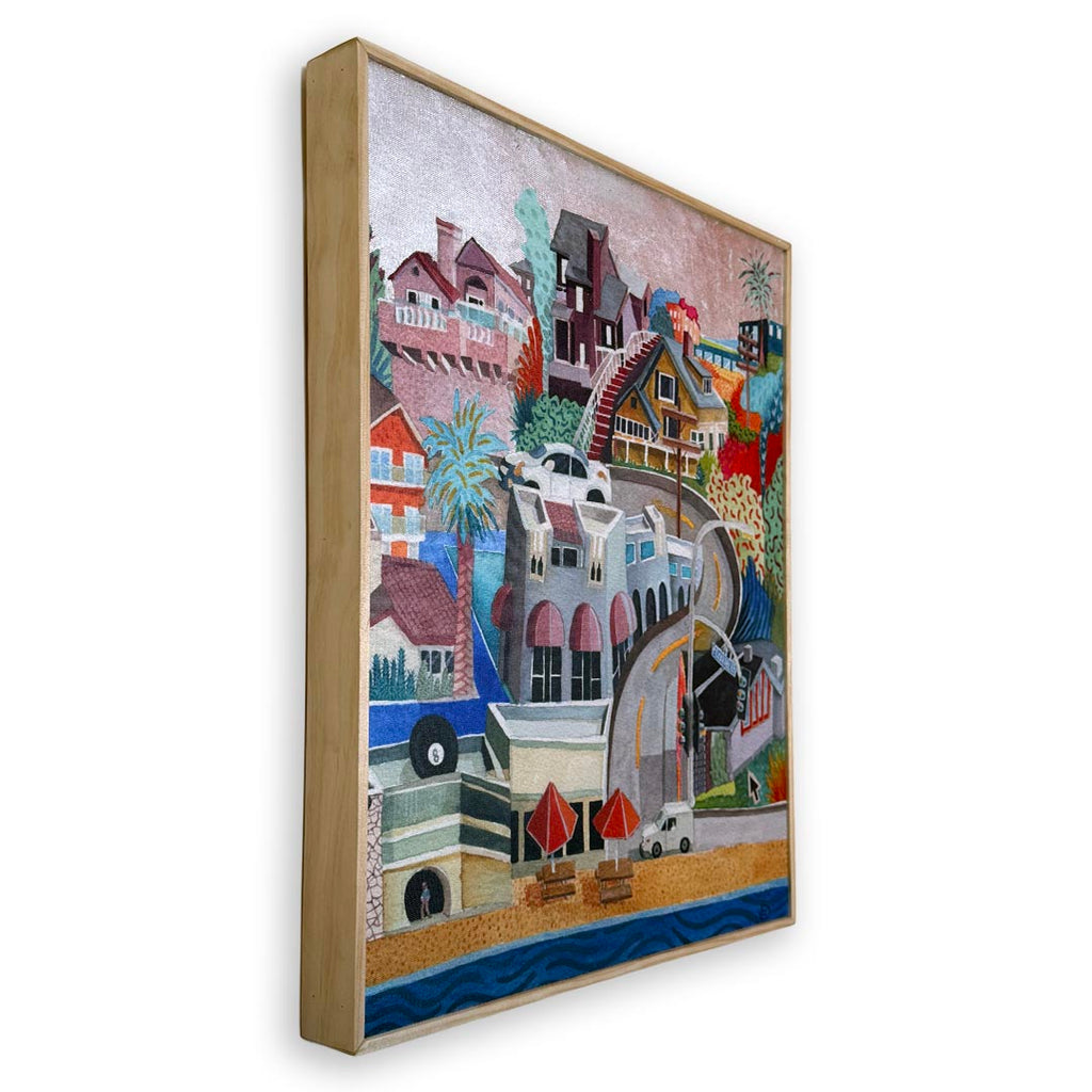 A framed mixed-media collage painting of a coastal town, shown at an angle against a white background. The artwork features stylized houses, palm trees, a beach, and a blue ocean, housed in a light natural wood frame. Silver foil sky.