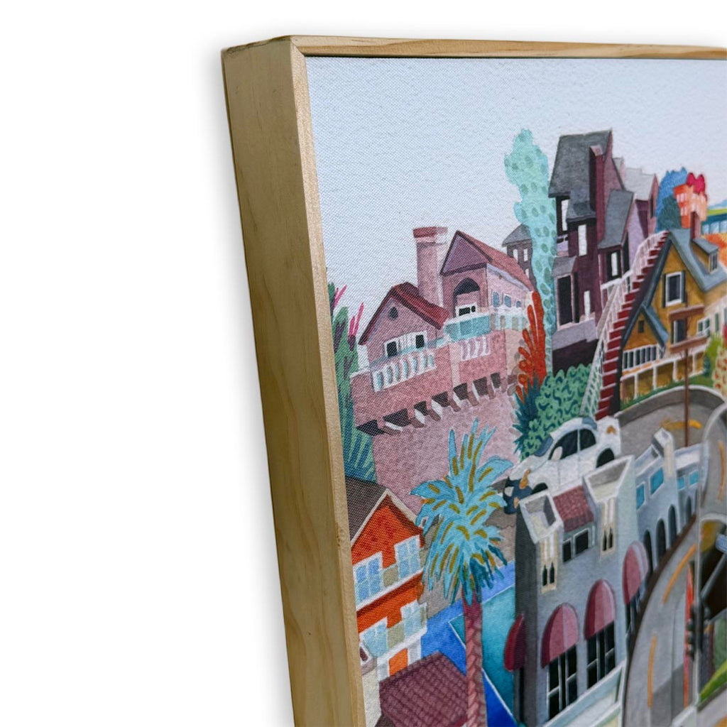 A close-up of a framed watercolor painting showing colorful houses and buildings on a hillside, with a light wood frame visible against a white background.