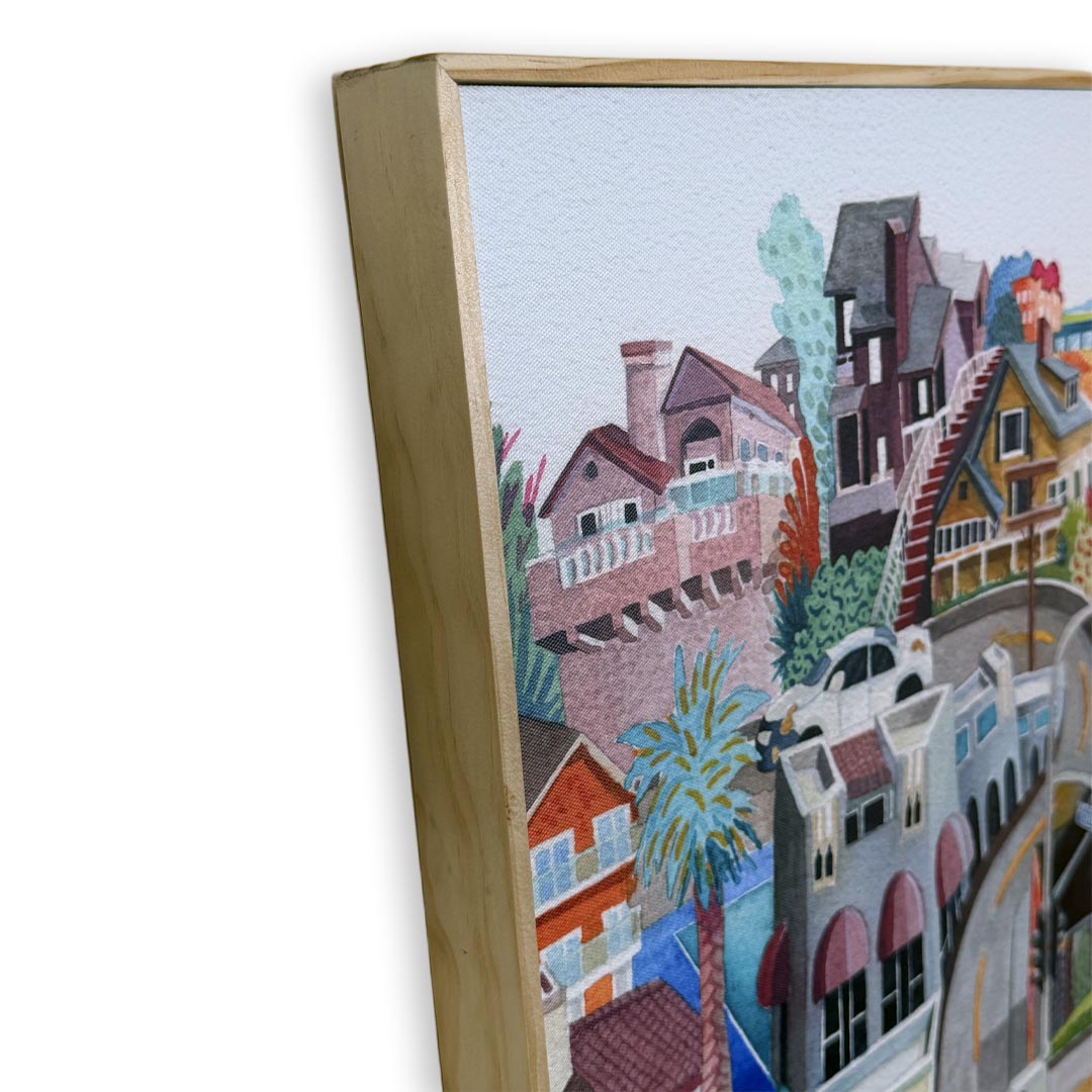 A close-up of a framed watercolor painting showing colorful houses and buildings on a hillside, with a light wood frame visible against a white background.