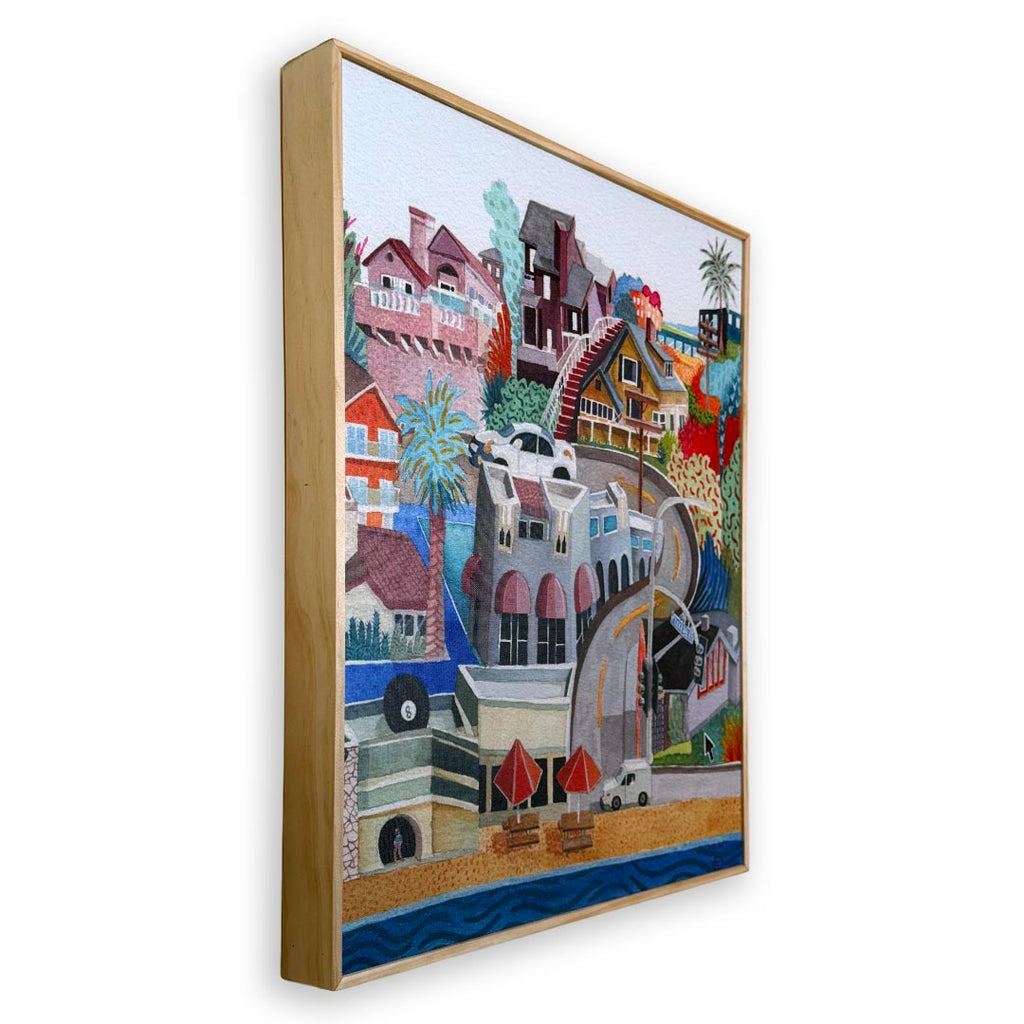 A framed canvas art print featuring a colorful, whimsical coastal town illustration with houses, palm trees, beach umbrellas, and blue ocean water, shown at an angle against a plain white background.