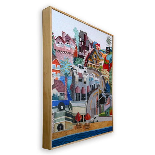 A framed canvas art print featuring a colorful, whimsical coastal town illustration with houses, palm trees, beach umbrellas, and blue ocean water, shown at an angle against a plain white background.
