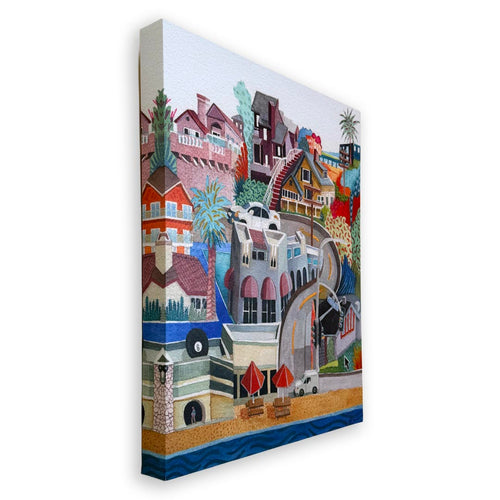 A colorful canvas wall art print depicting a whimsical coastal town scene with houses, palm trees, a lighthouse, beach umbrellas, and water, shown at an angle against a plain white background.